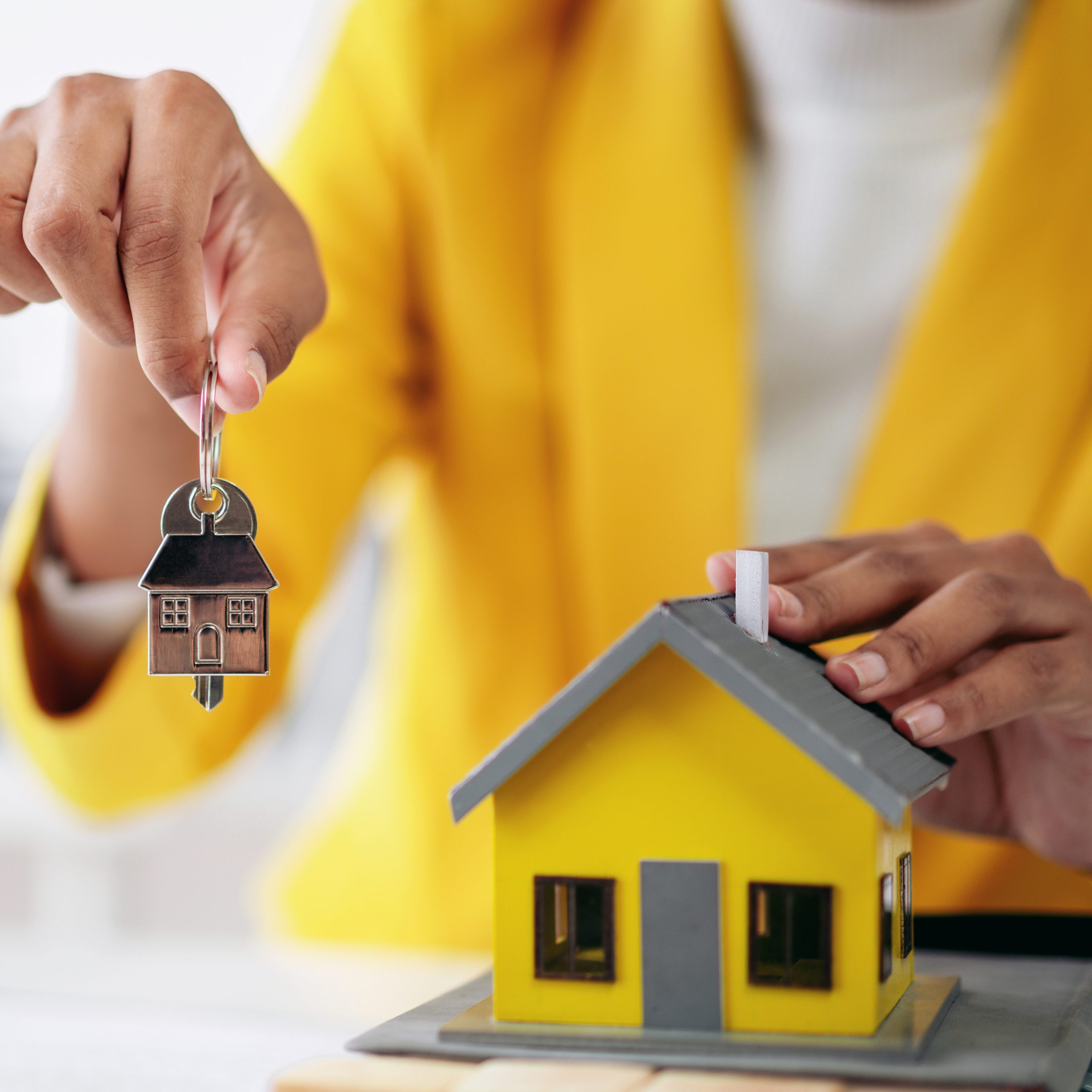 A woman is holding a key and yellow house. The house is a model and the woman is a real estate agent or a homeowner. Concept of loan and insurance.